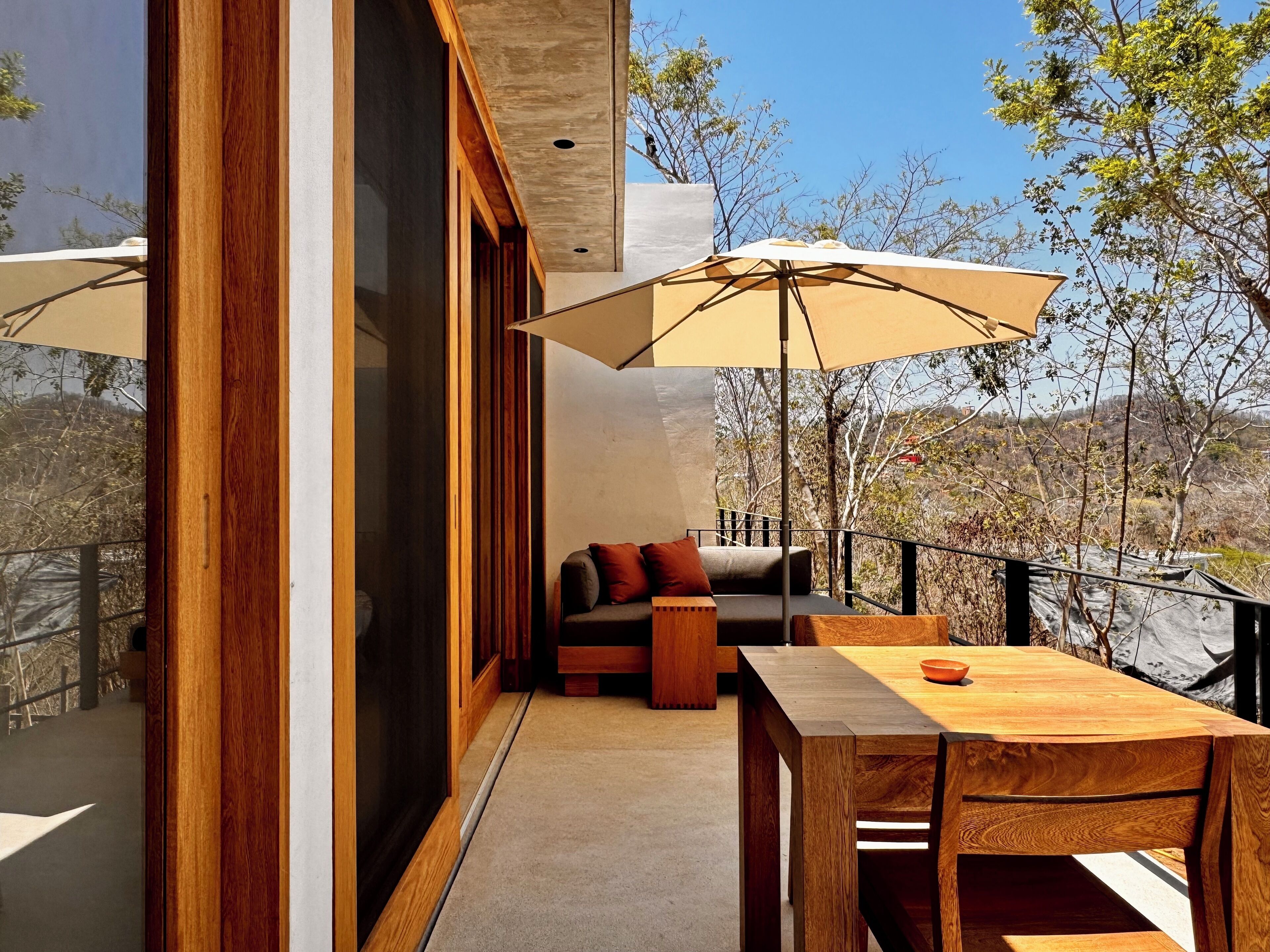 A modern outdoor seating area with a wooden table and chairs, a cushioned bench, and a large umbrella providing shade.
