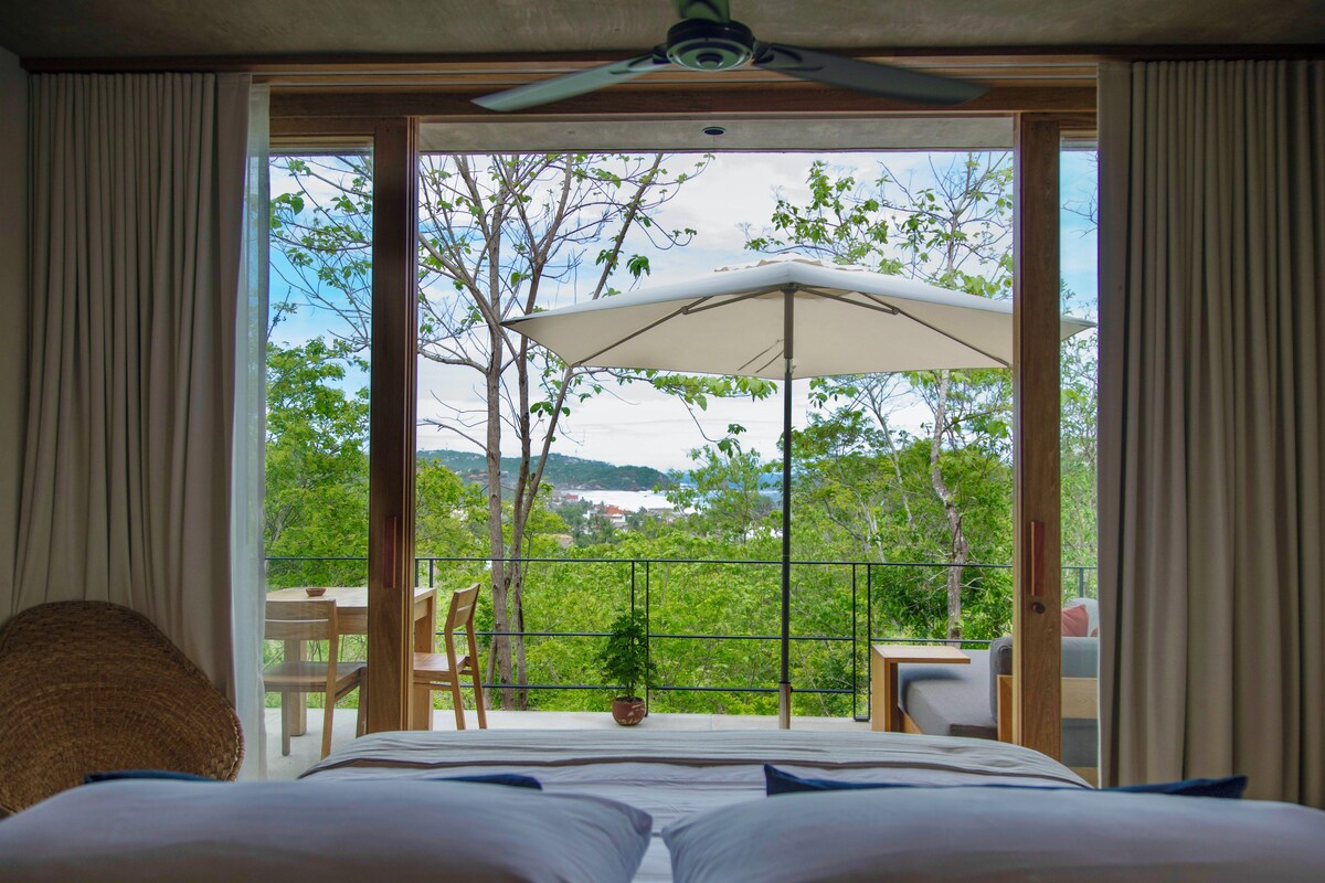 A bedroom with a large bed, a ceiling fan, and a view of a balcony with outdoor furniture and a potted plant.