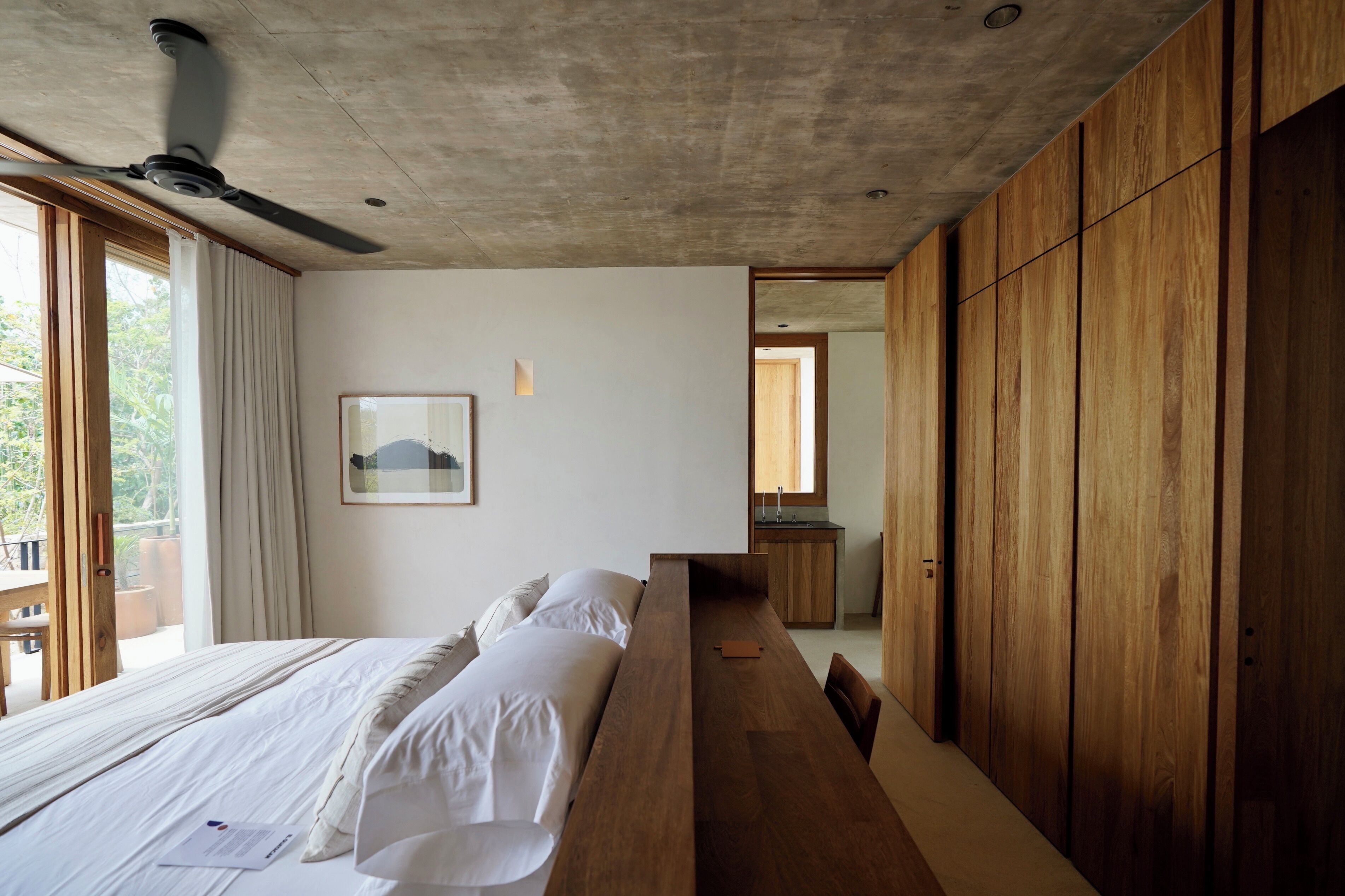 A modern bedroom with a large bed, wooden paneling, a ceiling fan, and a view of the outdoors.