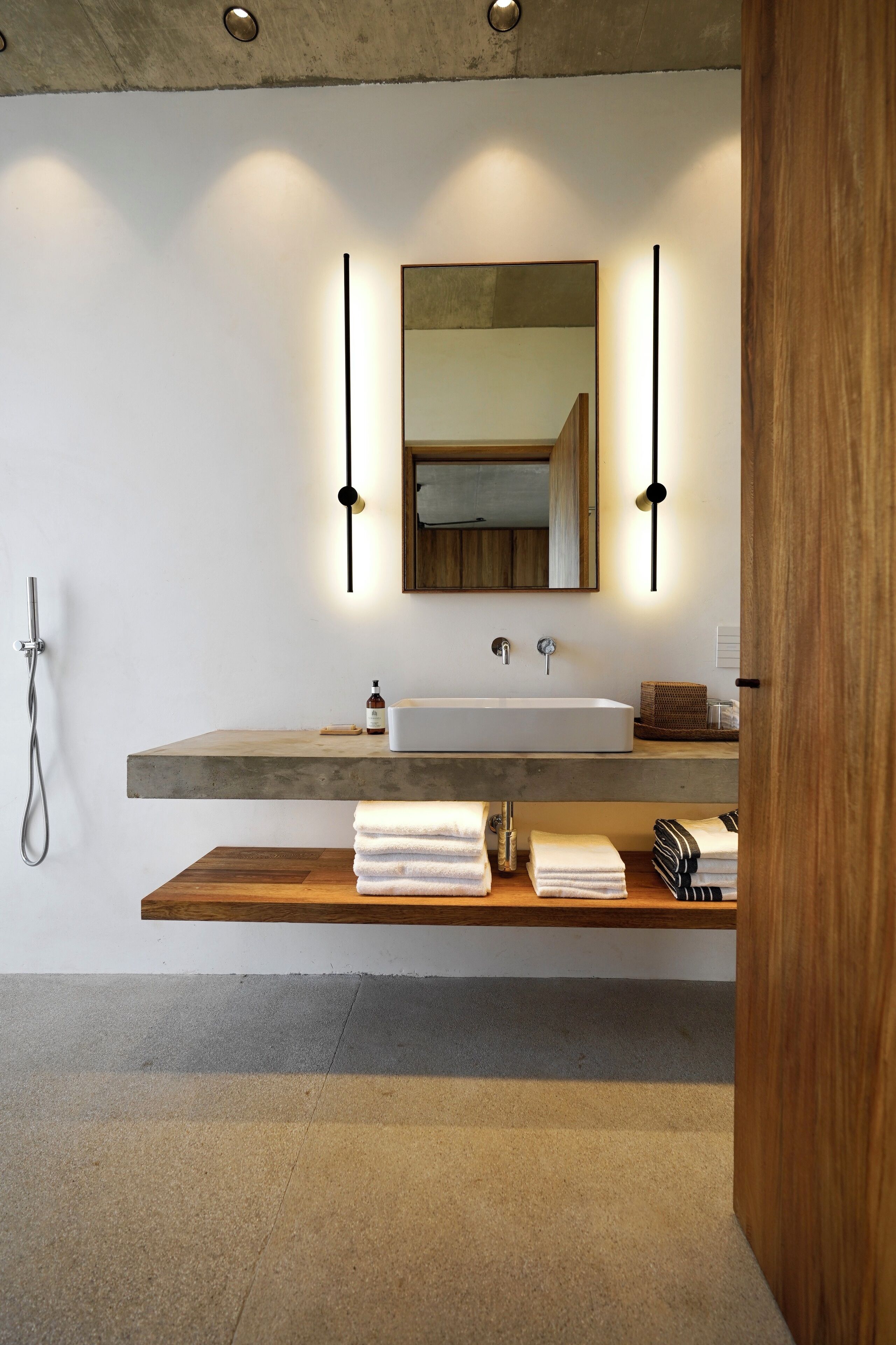 Une salle de bain moderne avec un grand lavabo, une étagère en bois où sont posées des serviettes, une douchette à main et un miroir avec des appliques murales.