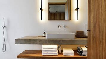 Un baño moderno con un lavabo grande, un estante de madera con toallas, un cabezal de ducha manual y un espejo con luces empotradas.