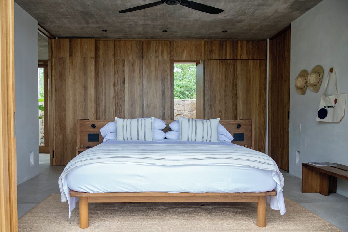 A bedroom with a wooden headboard, a large bed with striped bedding, a ceiling fan, and a stone wall.