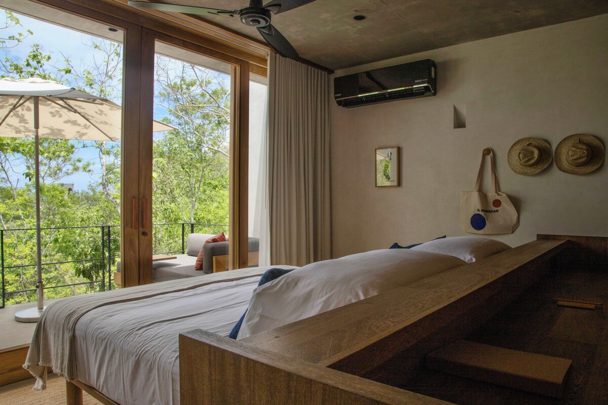 Un dormitorio con cama, ventilador de techo, ventana con cortinas y vista a la vegetación.