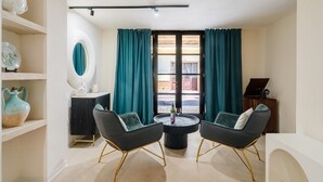 Una sala de estar moderna con una mesa redonda, dos sillas negras con patas doradas, cortinas color turquesa y vistas a través de una puerta de cristal.