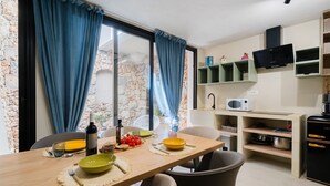 Un comedor con una mesa dispuesta para dos, una cocina con microondas y estanterías, y una pared de piedra visible a través de la ventana.