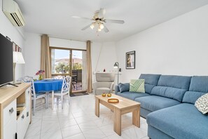 Un salon moderne avec un canapé bleu, une table basse en bois, une table à manger avec des chaises, un ventilateur de plafond et une grande fenêtre.