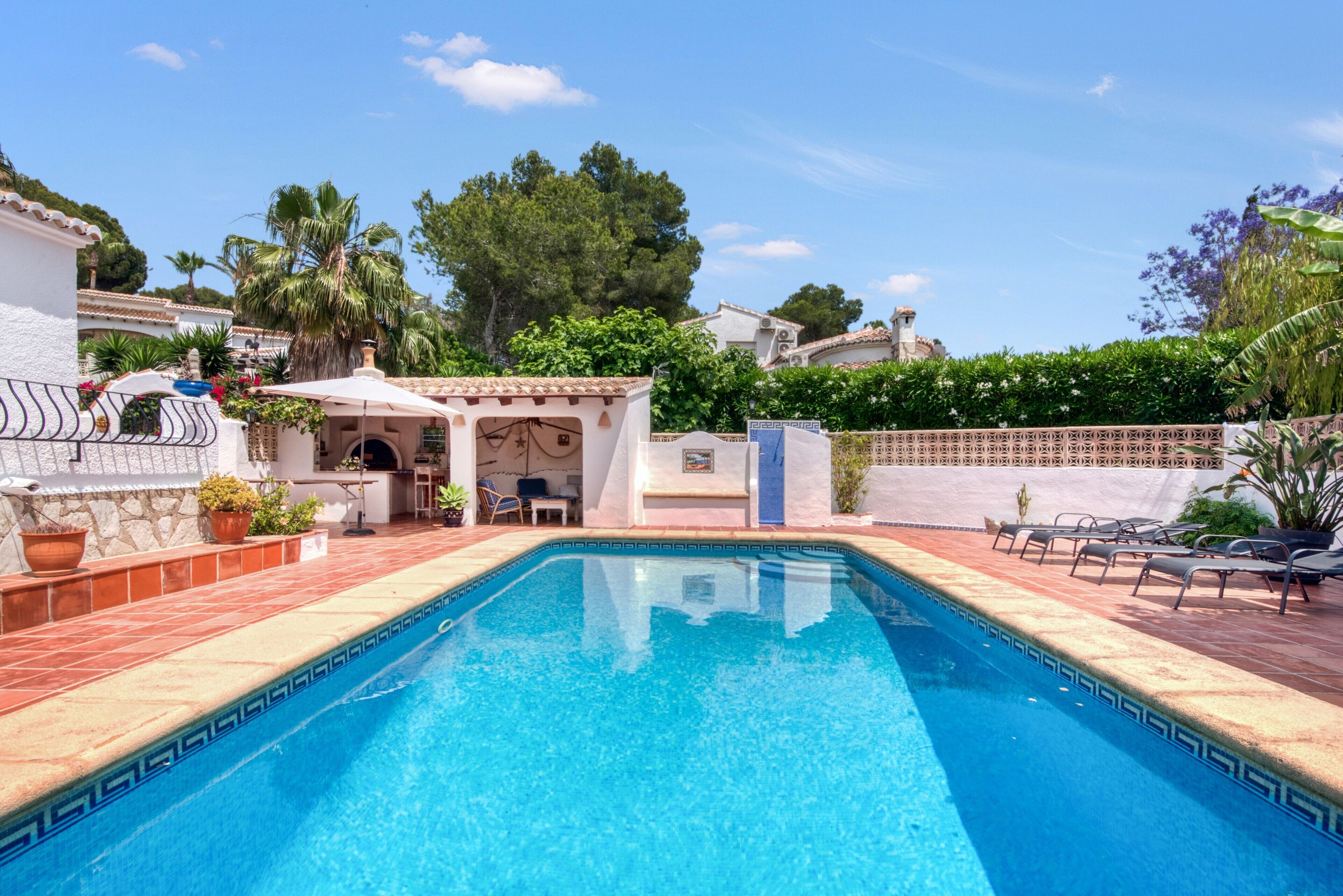 Une piscine dotée d’une terrasse carrelée, entourée de verdure et bordée d’un bâtiment blanc avec un patio.