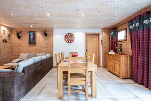 A wooden interior with a dining table, chairs, a sofa, a clock, and a kitchen area.