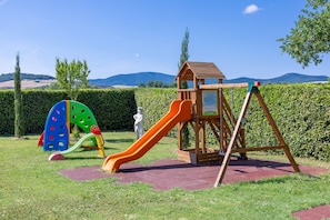 A playground with a slide and climbing structure, surrounded by a hedge and a view of hills.