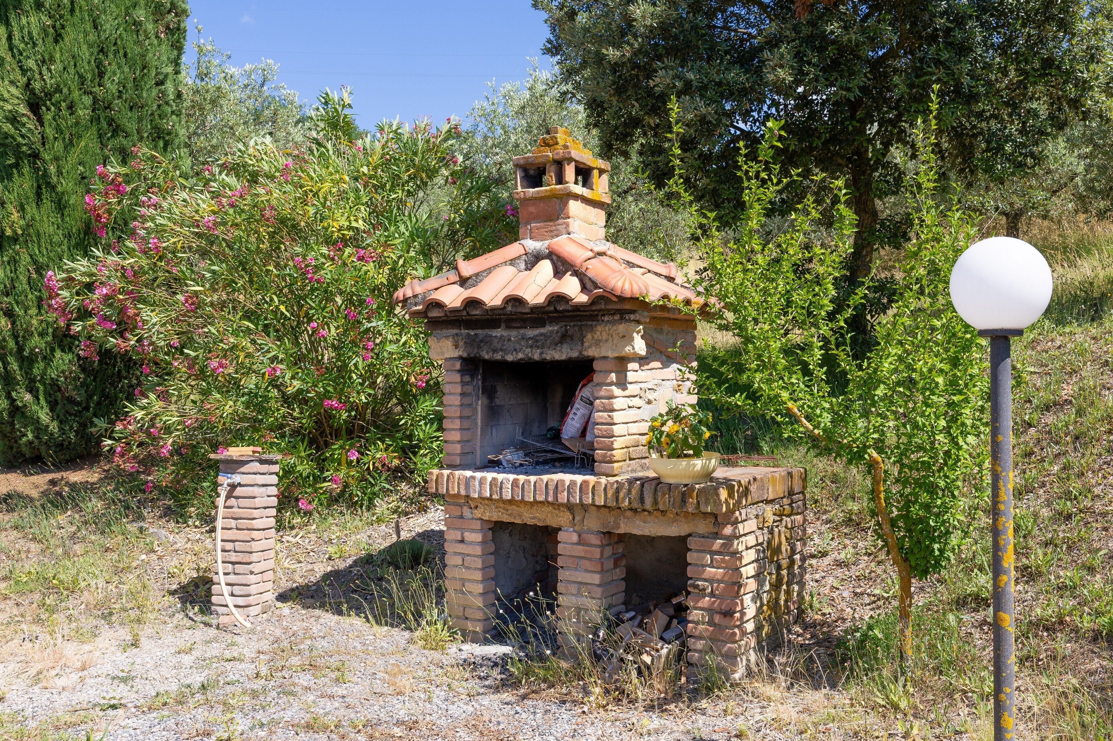 A brick-built outdoor oven with a chimney, surrounded by greenery and a lamp post.