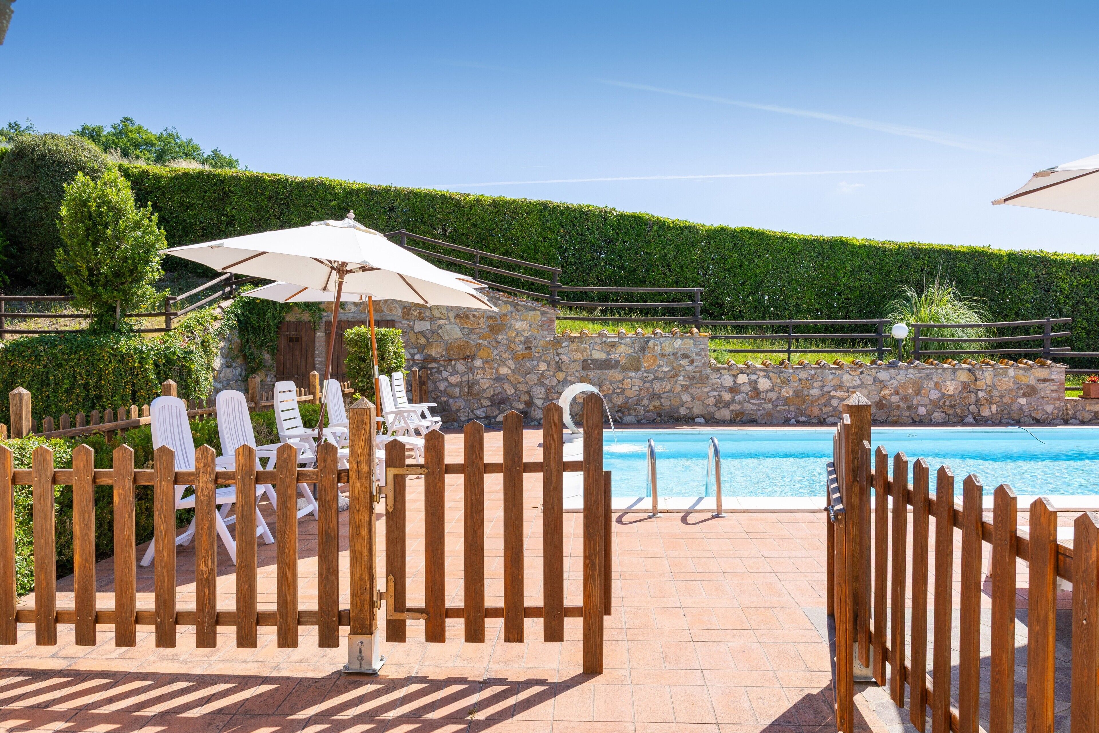 A pool area with a wooden fence, lounge chairs, and a stone wall.