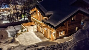 A wooden cabin with a snow-covered roof, illuminated windows, and a snow-covered driveway. - Chalet "Friwi": Mountain View, Private Terrace & Wi-Fi (Saint-Jean-de-Sixt)