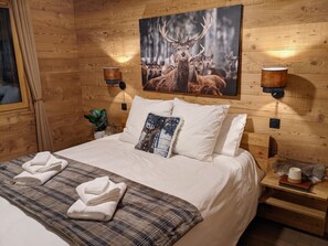 A wooden room with a bed, pillows, a blanket, towels, a wall-mounted lamp, and a framed picture of deer.