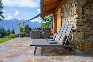 A stone patio with a lounge chair and cushions, a stone wall, and a mountain view.