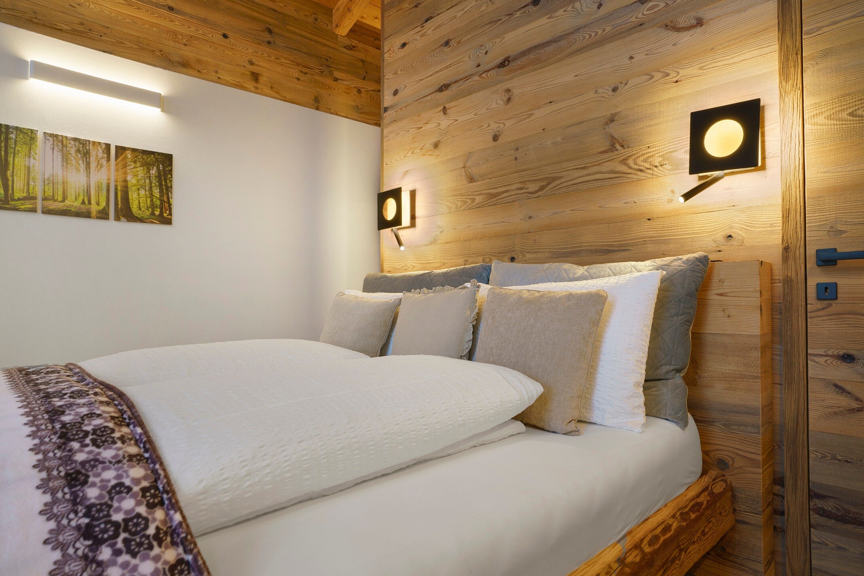 A wooden-paneled bedroom with a bed, wall-mounted light, and framed forest artwork.