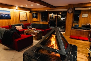 Ein gemütliches Zimmer mit Kamin, roten Sofas, einem Holztisch und Blick durch eine Glastür.