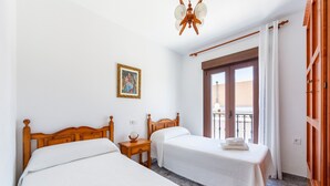 Ein Schlafzimmer mit zwei hölzernen Betten, einem Deckenventilator, einem kleinen Nachttisch mit Lampe, einem gerahmten Bild an der Wand und einer Balkontür.