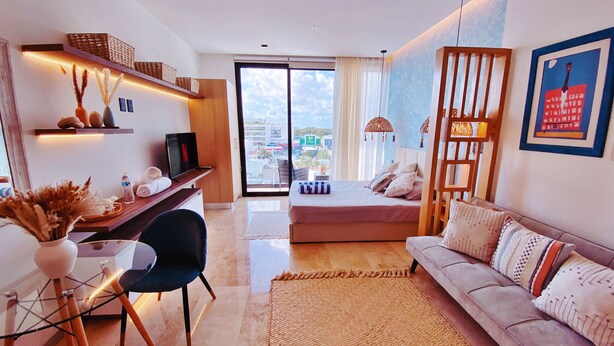 A modern living room with a sofa, a TV, a desk, and a view of the city through large windows. - Brand New Studio in 4th Floor with Balcony (Playa del Carmen)