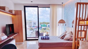 A modern bedroom with a bed, a desk with a TV, a chair, and a view of the city through a large window. - Brand New Studio in 4th Floor with Balcony (Playa del Carmen)