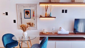 A modern living room with two blue velvet chairs, a glass coffee table, a wooden sideboard with decorative items, a flat-screen TV, and a framed artwork on the wall. - Brand New Studio in 4th Floor with Balcony (Playa del Carmen)