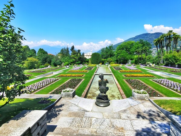 Un jardín formal con parterres simétricos, una fuente central y montañas distantes al fondo.