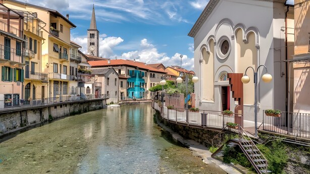 Ein Kanal mit Gebäuden an beiden Seiten, eine Kirche mit einem Spitzturm und eine blaue Fontäne.