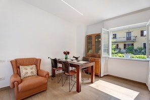 A dining area with a table, chairs, a leather armchair, a cabinet, and a window with a view of a building and greenery.
