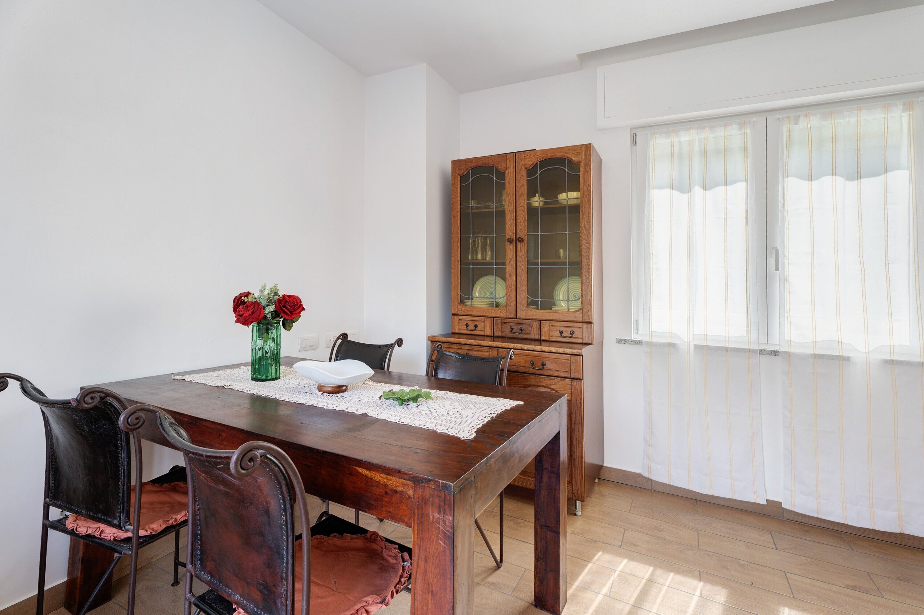Un comedor con una mesa de madera, sillas, un jarrón con flores, un aparador y una ventana con cortinas.