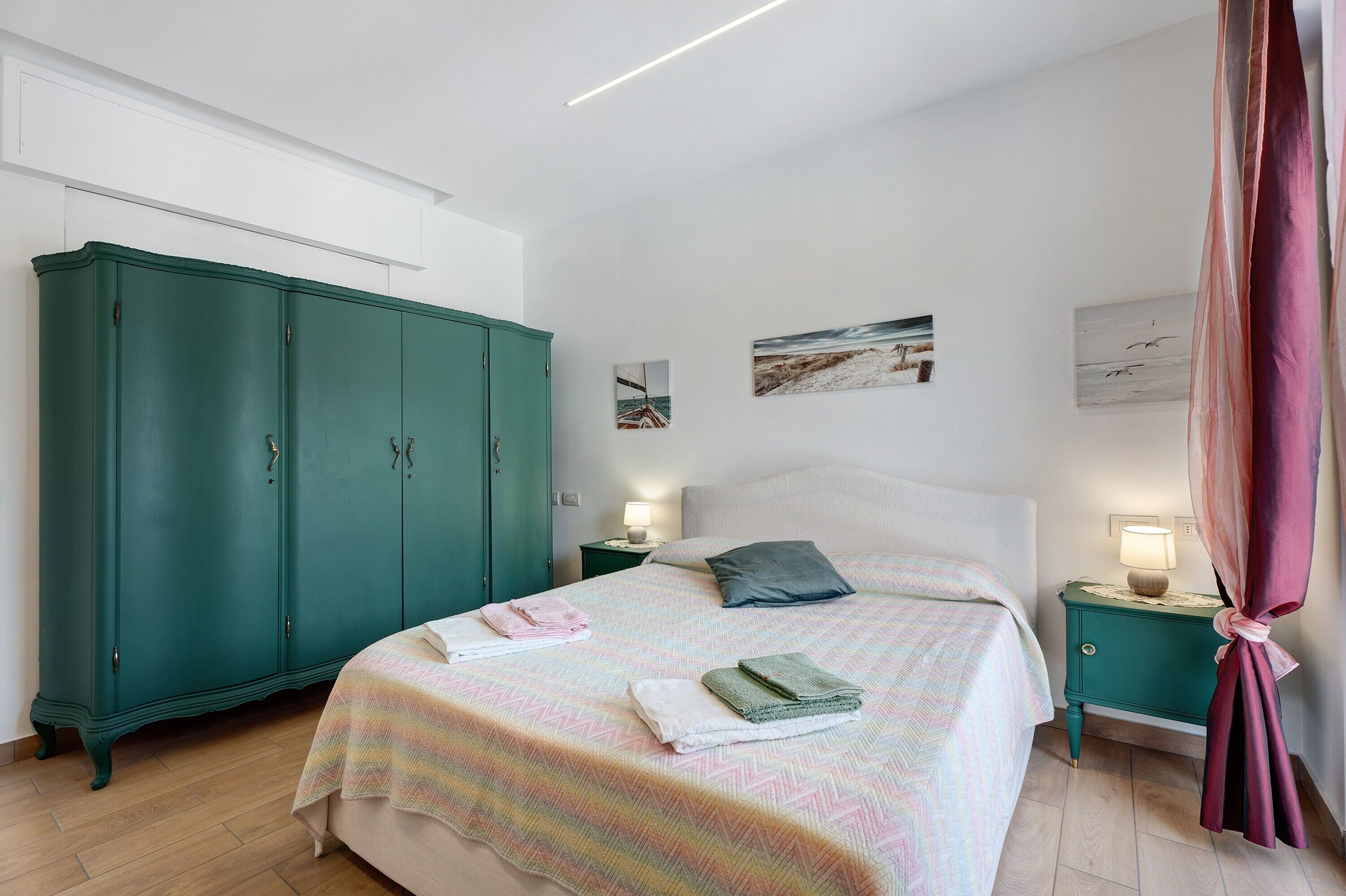 A bedroom with a bed, a green wardrobe, two bedside tables with lamps, and artwork on the wall.