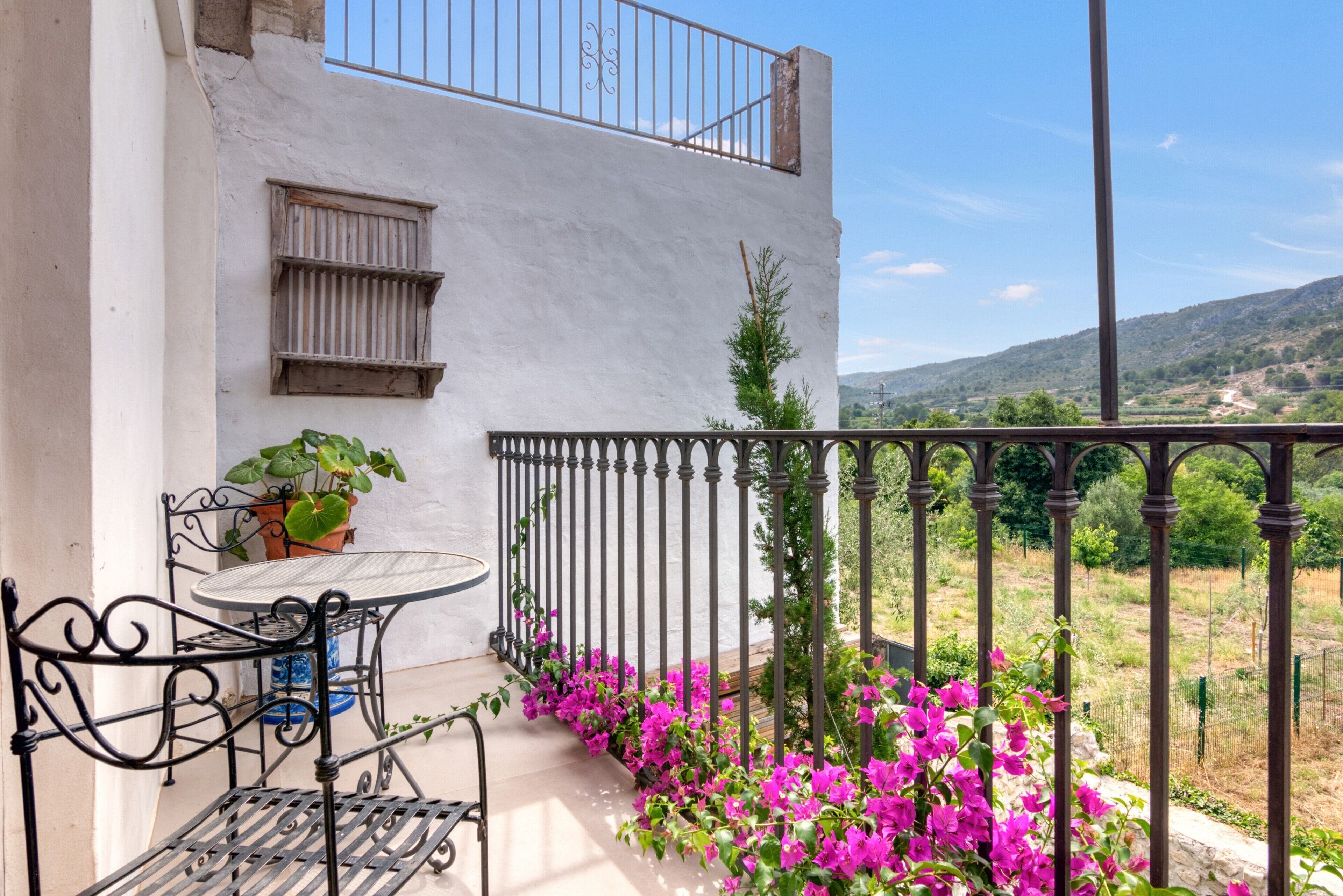 A balcony with a table and chairs, a railing, and a view of a hilly landscape.