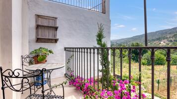 A balcony with a table and chairs, a railing, and a view of a hilly landscape.