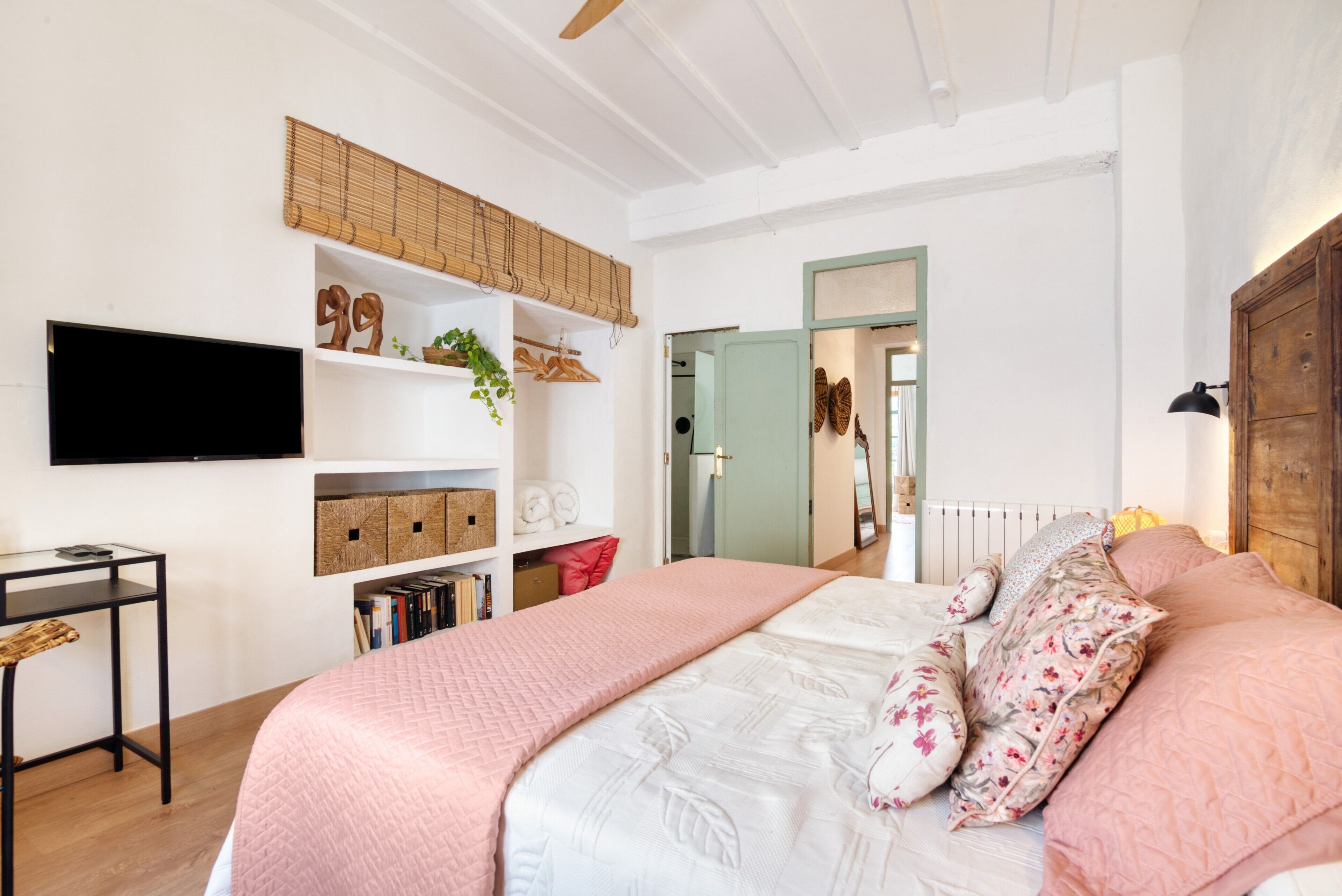 A bedroom with a bed, a TV mounted on the wall, a bookshelf, and a door leading to another room.