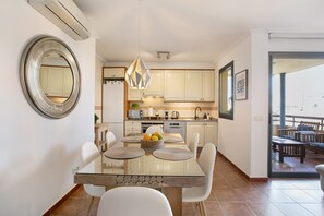 Una cucina moderna con mobili bianchi, una zona pranzo con tavolo in vetro e sedie bianche, e vista attraverso una porta vetrata su un'area relax all'aperto.