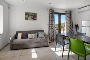 Ein zeitgemäßer Wohnraum mit einem grauen Sofa, einem Esstisch und einem grünen Stuhl. Ein Bild ist an der Wand zu sehen, und ein Fenster mit Vorhängen ist erkennbar.