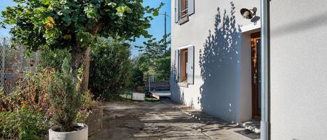 A narrow pathway leading to a building with a white exterior, a wooden door, and a potted plant on the left.