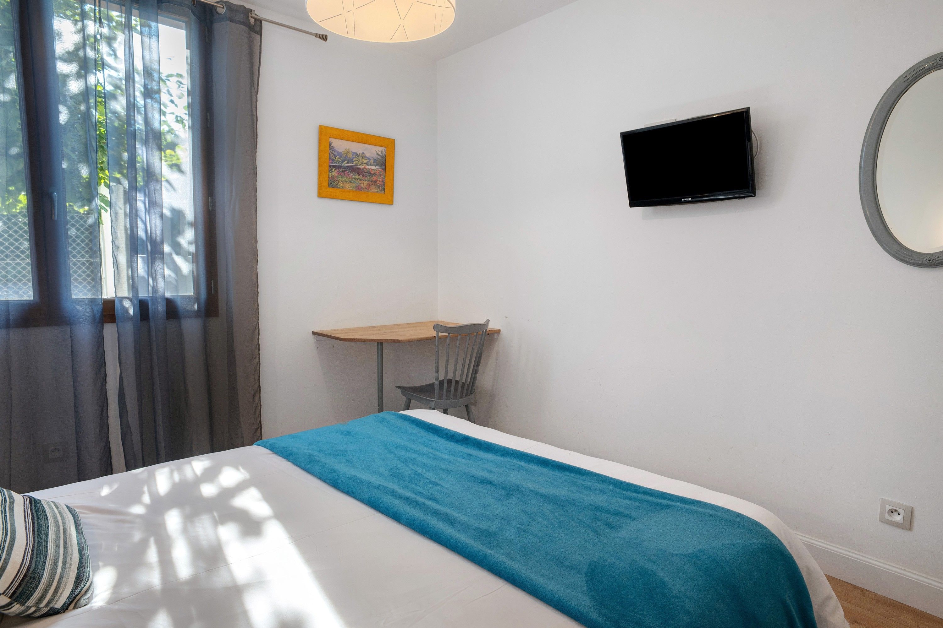 Een slaapkamer met een bed, een bureau, een stoel, een televisie en een raam met gordijnen.