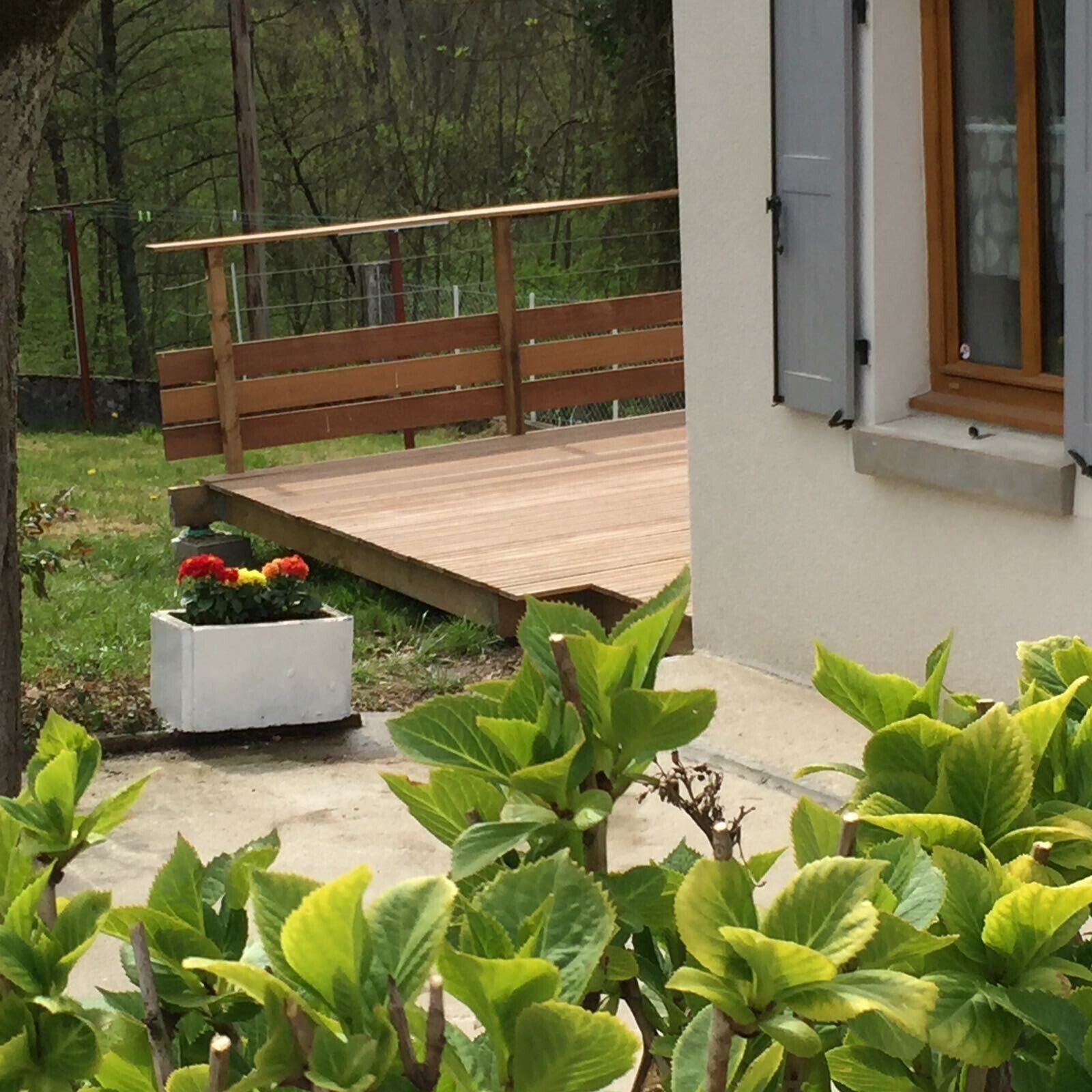 Une terrasse en bois, un jardinier blanc orné de fleurs et des plantes vertes dans un espace aménagé en jardin.