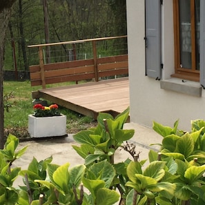 En træterrasse, en hvid plantekasse med blomster, og grønne planter i en have.