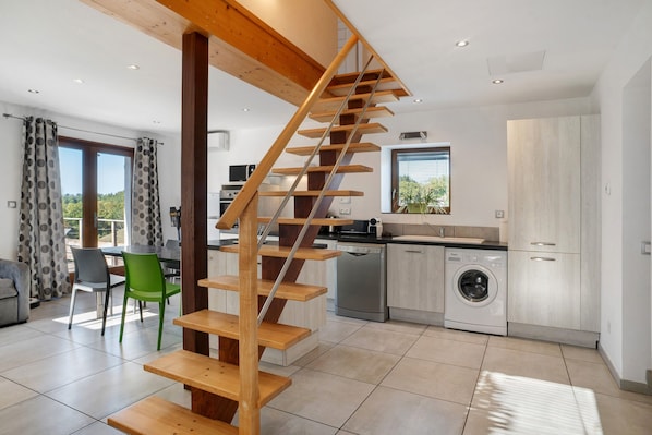 A modern kitchen with a built-in washing machine, a dining area with a green chair, and a wooden staircase.