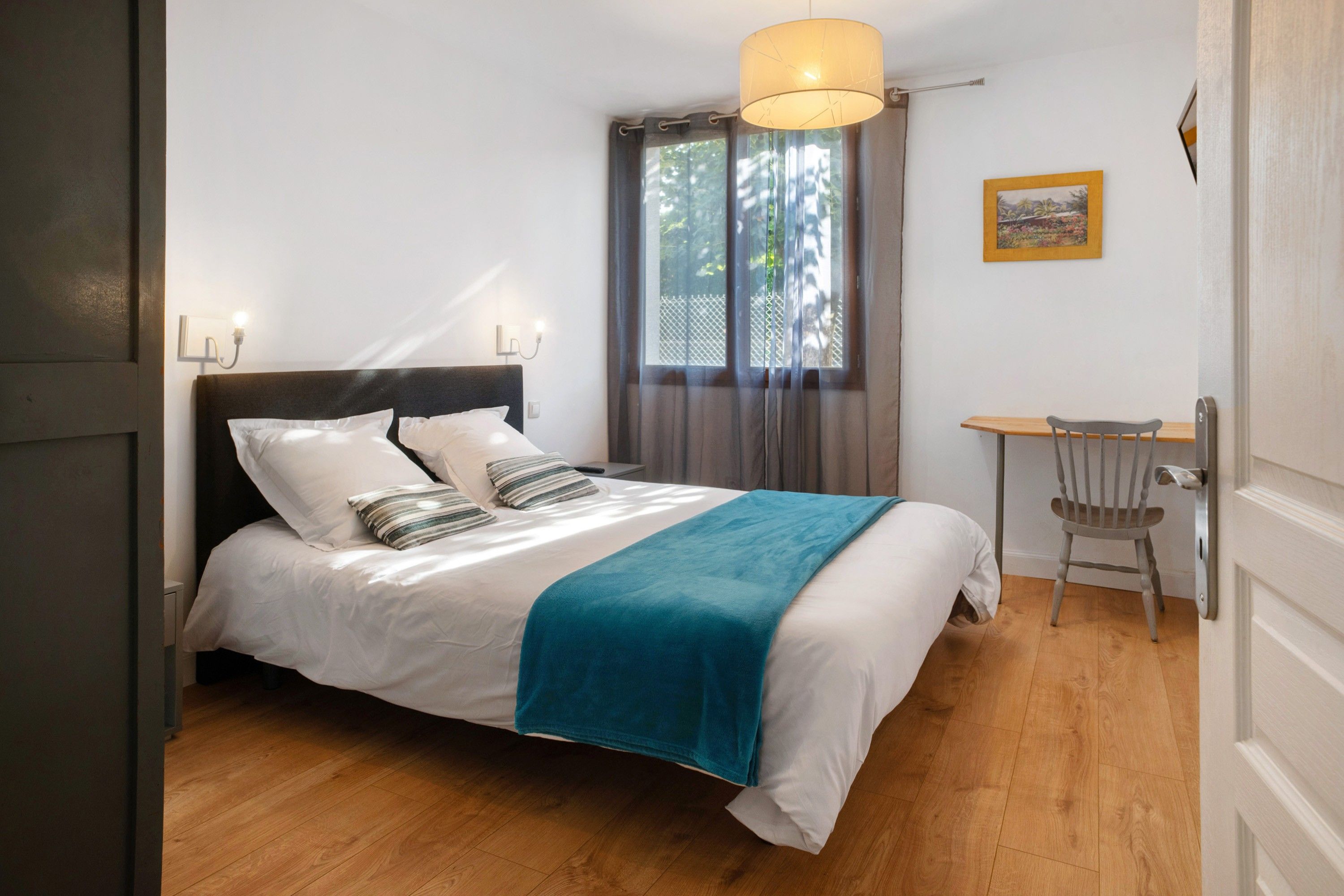 Una cama bien hecha con un plaid verde azulado, una silla de madera, una mesita y una ventana con cortinas.