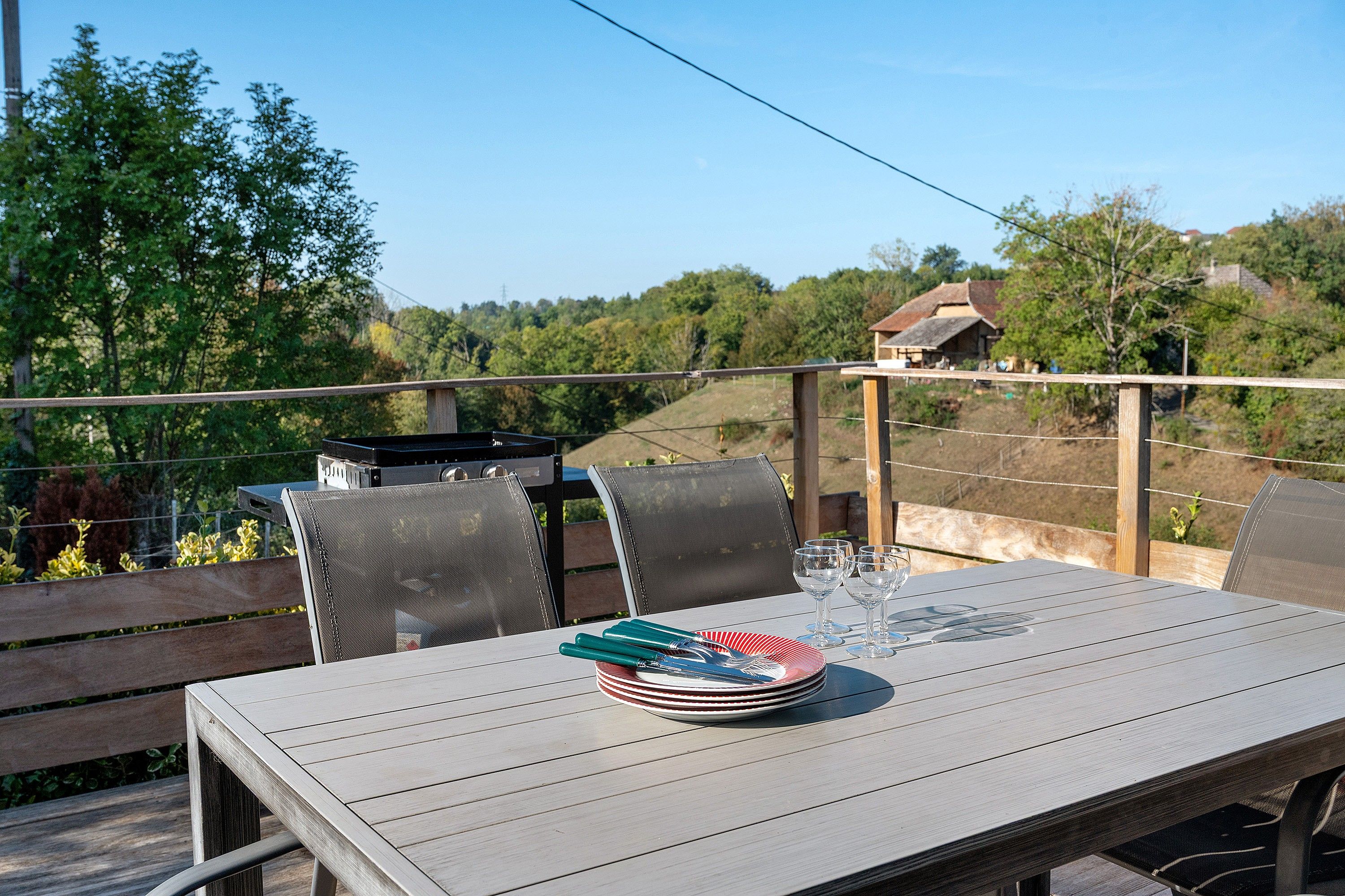 Un área de comedor al aire libre con una mesa preparada para cuatro personas, rodeada de sillas, y con vistas a un paisaje pintoresco con árboles y una casa al fondo.