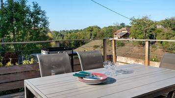 Un área de comedor al aire libre con una mesa preparada para cuatro personas, rodeada de sillas, y con vistas a un paisaje pintoresco con árboles y una casa al fondo.