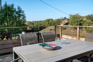 Un área de comedor al aire libre con una mesa preparada para cuatro personas, rodeada de sillas, y con vistas a un paisaje pintoresco con árboles y una casa al fondo.