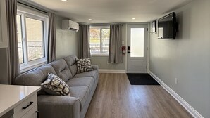 A modern living room with a sectional sofa, a flat-screen TV mounted on the wall, and a kitchen area with a white countertop and cabinets. - ORS Beach Corporation | Wasaga Beach Rental (Wasaga Beach)