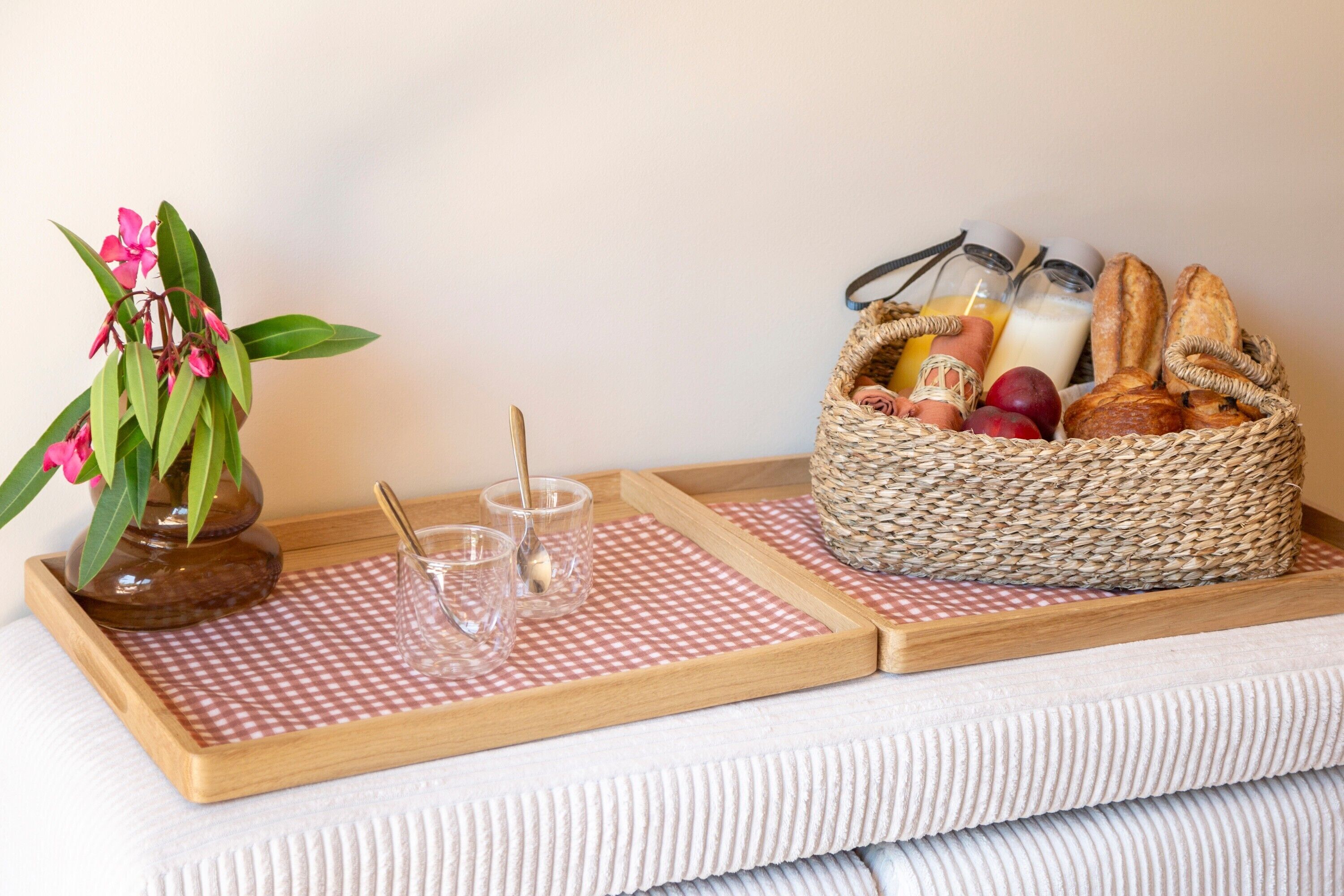 A tray with a checkered tablecloth, a vase with pink flowers, two glasses with spoons, and a basket containing bread, apples, and bottles.