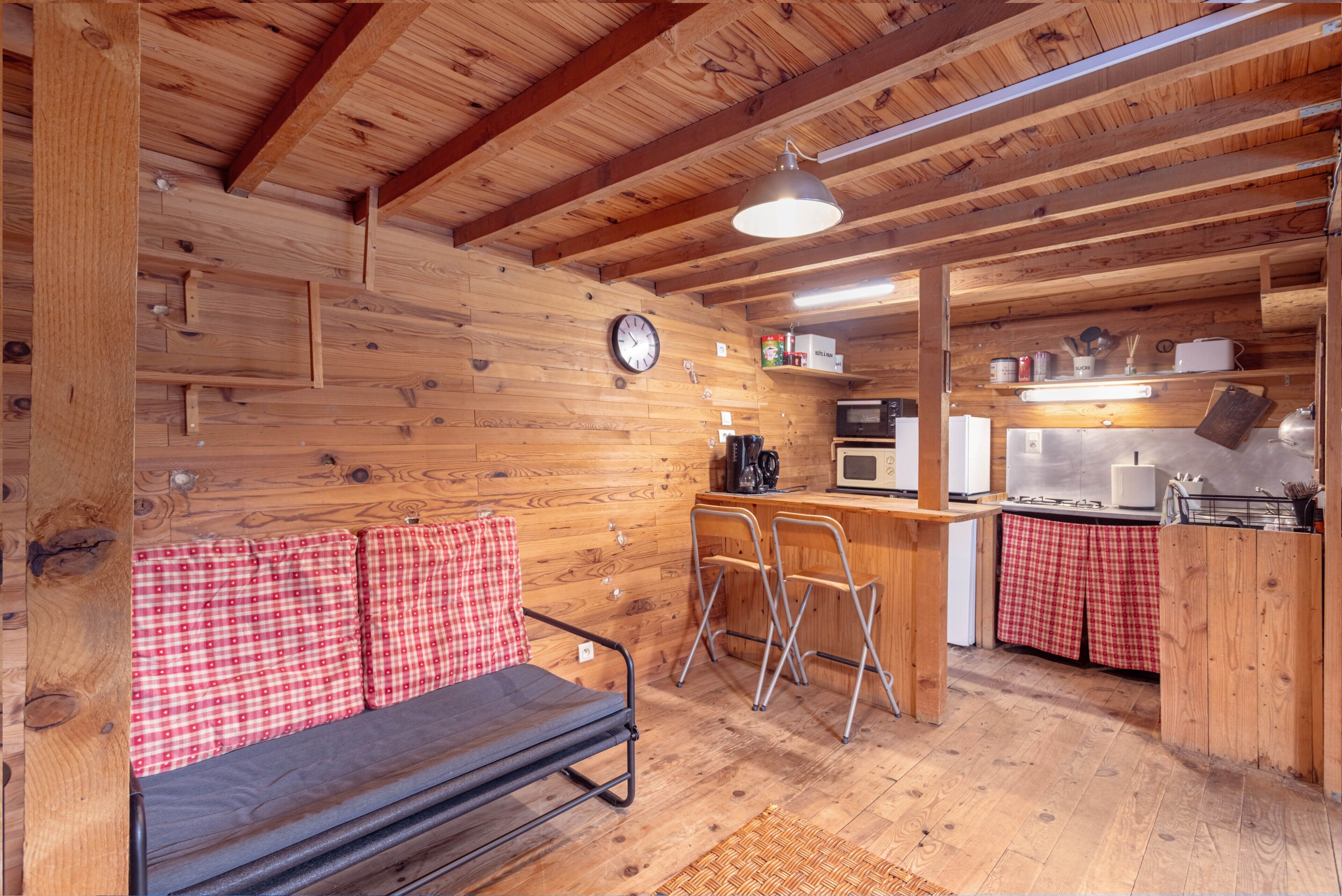 Un accogliente interno di una baita con soffitto in legno, un divano con coprimuri a quadri e un angolo cottura con microonde, fornello e sgabelli da bar.