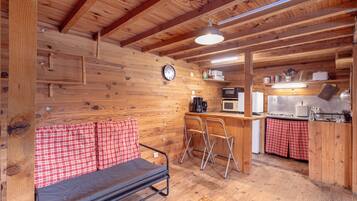 Un accogliente interno di una baita con soffitto in legno, un divano con coprimuri a quadri e un angolo cottura con microonde, fornello e sgabelli da bar.