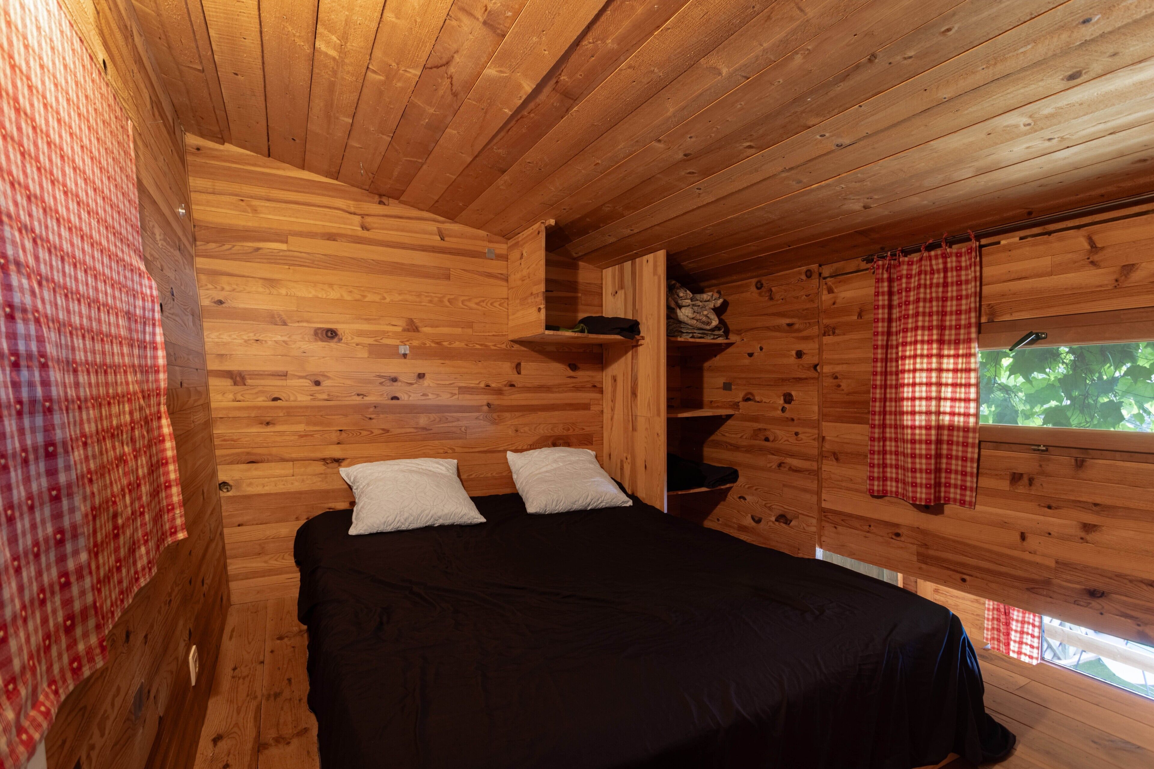 Quarto de cabana de madeira com cama, almofadas, prateleira e janela com cortina xadrez.