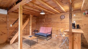 A wooden cabin interior with a bed, a table, a chair, a clock, and a shelf with items.