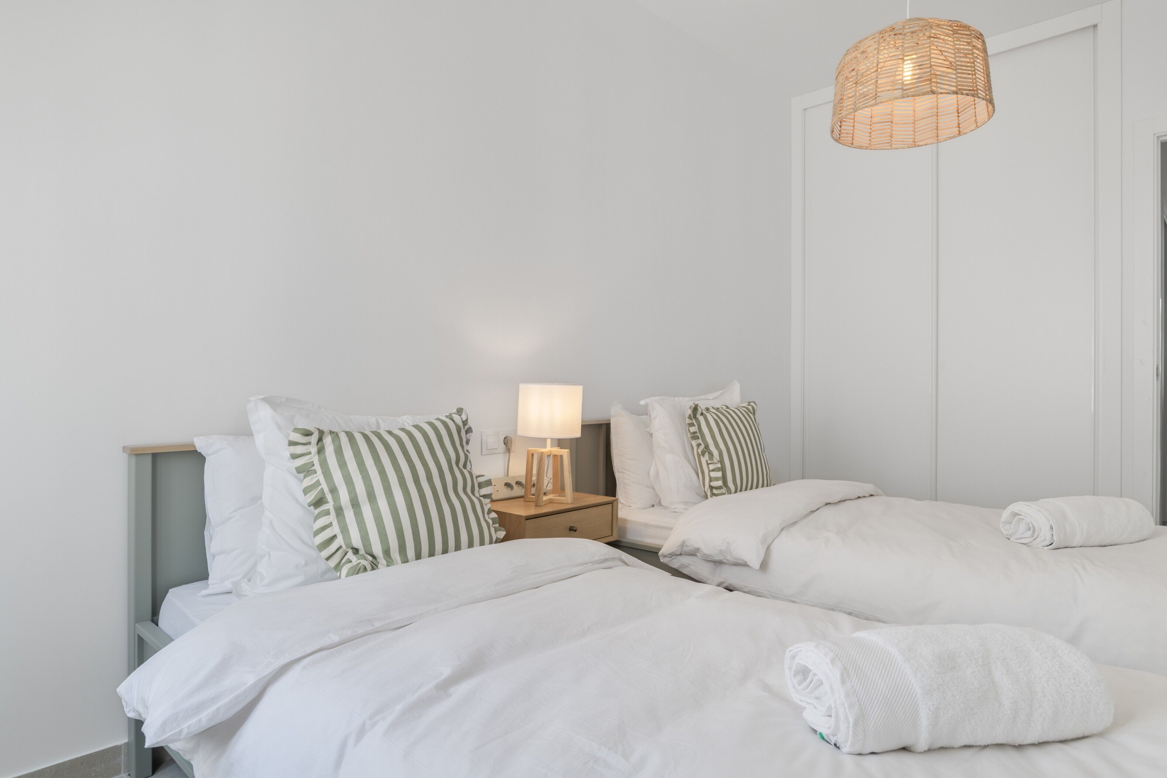 A hotel room with two beds, a bedside table with a lamp, and a wicker hanging light fixture.